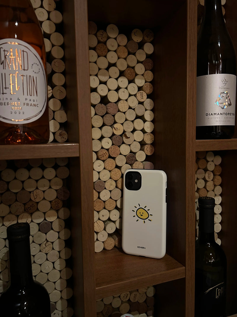 Dizajnový kryt na iPhone Kovera s ilustráciou, mobil vystavený na polici medzi fľašami vína, reálne použitie produktu